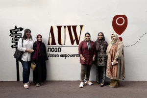 Five young Southeast Asian women, most of whom are wearing hijab, stand in front of a wall painted with the words AUW, noun, women empowerment.
