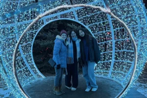 Three young women wearing winter jackets stand inside a blue glowing snow globe at a holiday lights display.