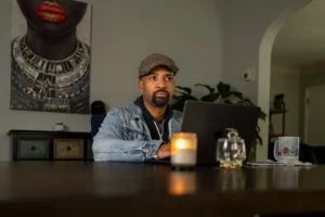 A Black man wearing a jean jacket and a newsboy hat sits at a table with a laptop and a candle.