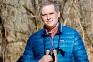 A headshot of a middle-aged white man with gray hair. He is wearing a blue puffy jacket and has binoculars on a string around his neck. He stands in a wooded area with bare tree branches in the background.