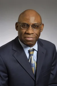 A headshot of a middle-aged Black man. He is bald and wears glasses. He is wearing a black suit with a blue shirt and tie. 