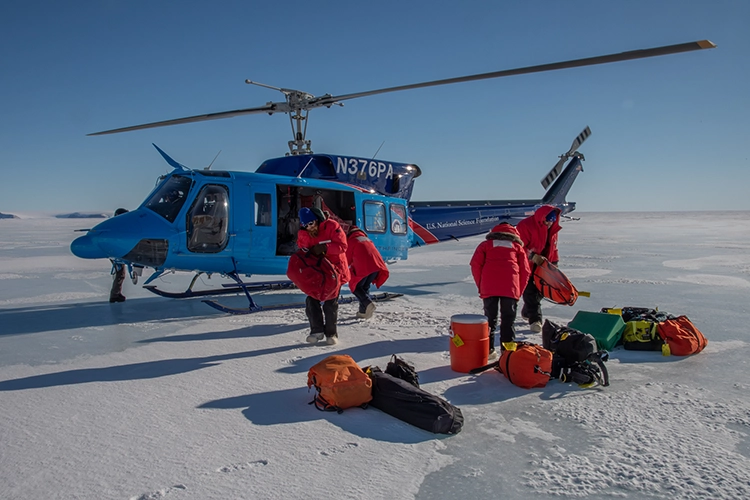 Two people in orange coats unload orange gear bags from a blue helicopter parked on a sheet of white ice.