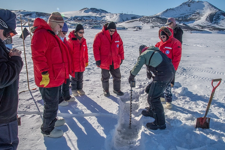 A group of people wearing orange cold-weather coats and gear stand around a man using an auger to drill a hole into ice. They are in a snowy landscape with snowy mountains in the distance.