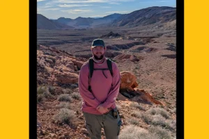 A young white man in a long-sleeved shirt and cargo pants stands in a dry, rocky landscape with mountains in the background.