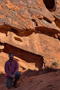 A young white man in a long sleeve shirt and cargo pants kneels in front of a red stone cliff wall. Behind him are petroglyphs etched in the rock depicting animals like horned sheep.