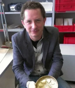 A middle-aged white man sits in an office, holding a golden cup in his hands.