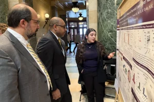 A young woman with dark long hair wearing business casual gestures to her research poster while two old men in suits look at her work.