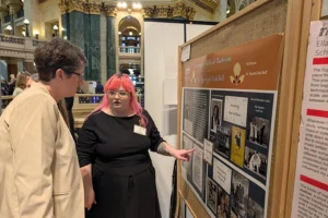 A young person with long pink hair wearing a black blouse gestures to a research poster while an older white woman in a blazer and glasses looks on.