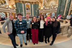 Six students in business casual dress stand in a line with their adult researcher mentor to one side. They are in the Wisconsin Capitol Rotunda, with intricate architectural columns in the background.