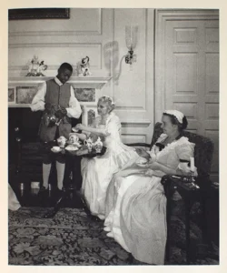 A historical, black-and-white image of a parlor in Colonial Williamsburg historical village where two white ladies in colonial dresses sit while a Black man dressed as a servant pours tea.