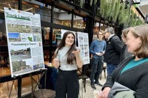 A young white woman with long dark hair gestures to a poster as she speaks with an older white woman. They are standing outside under an overhang with trailing ivy.