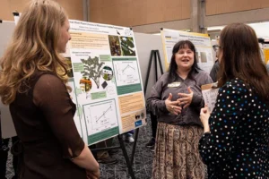 A white woman wearing a gray button-down shirt and skirt talks stands in front of a research poster and speaks to two women with their backs to the viewer.