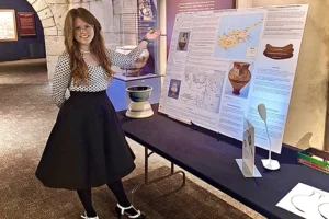 A young white woman with long brown hair and glasses wearing a black skirt stands at a table in front of poster showing a clay pot and other ancient artifacts.