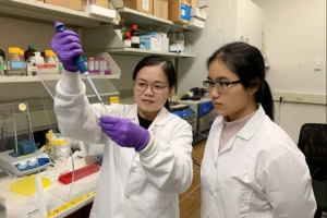 Two Asian women in white lab coats stand next to each other. The older woman on the left holds up a test tube and pipette as the younger woman on the right looks on.