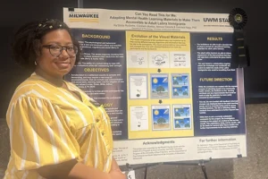 A Black woman with glasses and a yellow-striped blouse stands next to a research poster.