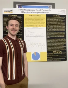 A young white man with brown hair and mustache wearing a maroon striped shirt stands in front of a yellow and black research poster.