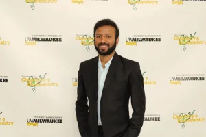 A young Indian man wearing a white button-down shirt and a black blazer stands in front of a yellow background with a repeating UW-Milwaukee logo.