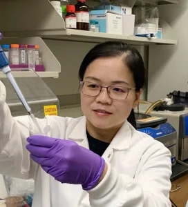 An Asian woman with dark hair and wearing glasses, purple gloves, and a white lab coat, pipettes a liquid into a container.
