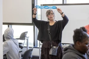 A middle-aged white woman holds up a line of blue paper chain links at the head of a classroom.