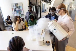 A white woman wearing safety goggles pours a billowing cloud of liquid nitrogen into a container as several middle school students look on.