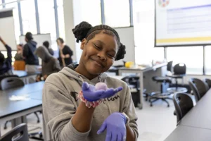 A young Black girl wearing purple latex gloves holds up a pale purple bouncy ball in her palm.