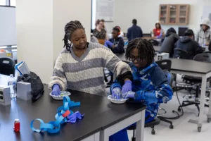 Two young Black students wearing purple latex gloves stir a glue mixture in a paper bowl with their fingers.