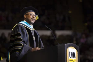 A Black man in graduation regalia stands at a podium marked with the UWM logo.