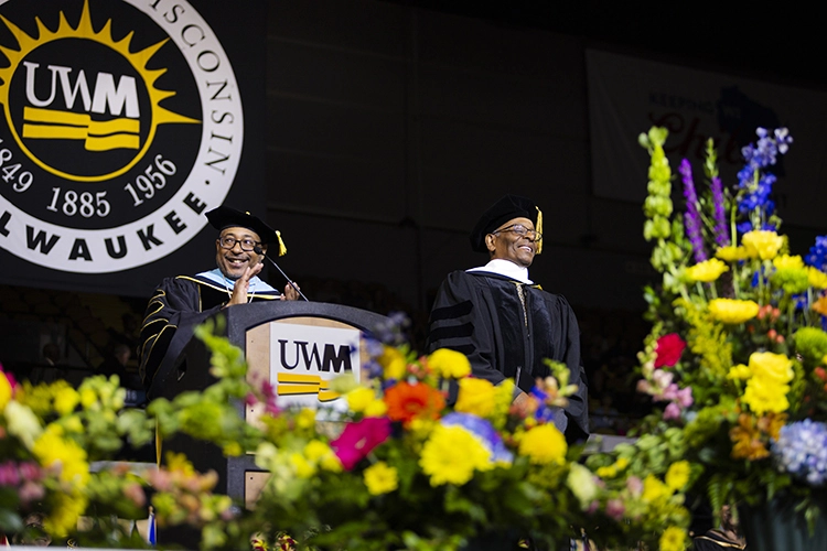 Two Black men, both wearing graduation regalia, stand on stage in front of the UWM seal with flowers in the foreground.