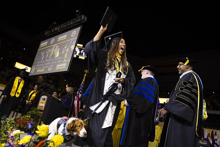 A young woman in a graduation cap and gown holds a diploma aloft with a huge smile. She holds the leash of a brown and white service dog.