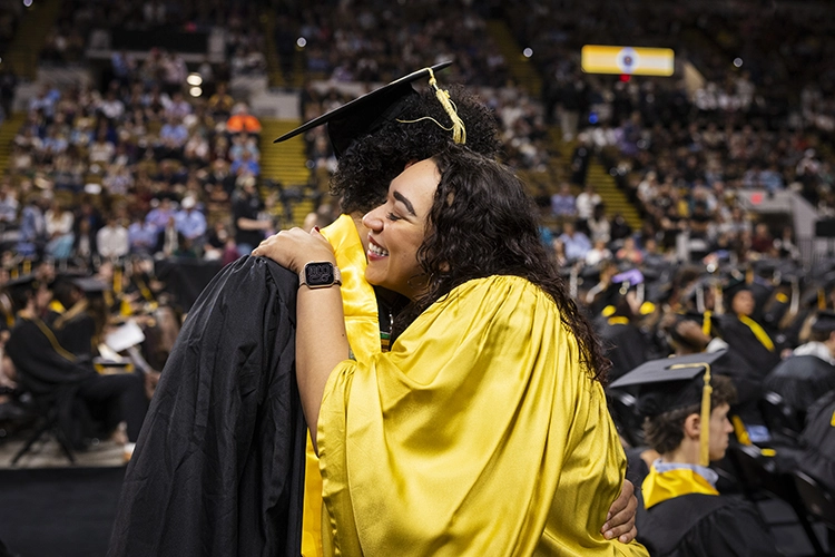 A Latina woman with curly dark hair wearing a gold graduation robe embraces a young man wearing a black robe and mortarboard.