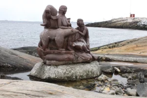 A red stone statue of a woman surrounded by a walrus, polar bear, and person, and other animals sits in front of an ocean shoreline.