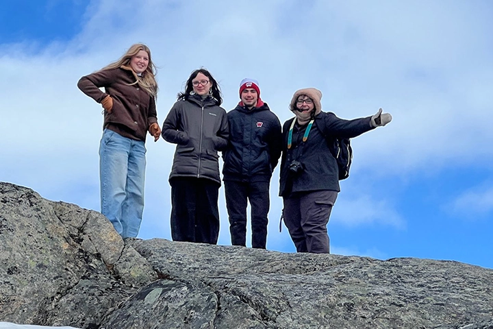 A group of four white students dressed in winter coats and hats stand on a gray rock, silhouetted against a blue sky with light white clouds.