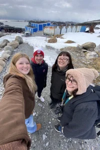 Four white students in winter coats take a selfie while standing on a rocky, snowy path with blue buildings in the background.