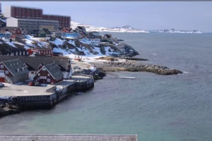 The shoreline of Nuuk in Greenland. Colorful buildings sit atop a snowy ridge next to the ocean.