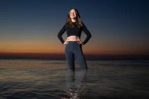 A white woman with long brown hair wearing a black cropped shirt and leggings stands-knee deep in water with the sky darkening at sunset behind her.