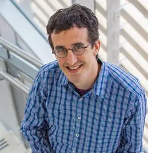 A head shot of a middle-aged white man with short, slightly curly black hair. He wears black glasses with round frames and a blue plaid, collared button-down shirt.