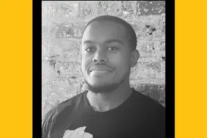 A black-and-white headshot of a young Black man with short hair and a beard on his chin, standing in front of a brick wall.