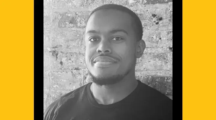 A black-and-white headshot of a young Black man with short hair and a beard on his chin, standing in front of a brick wall.