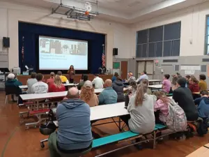 A group of people sits at several cafeteria tables in a school gym facing a projector screen. A woman stands before the screen with a microphone in hand.