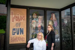 Two middle-aged white woman standing together at a glass bus shelter. Posters on the walls of the shelter read Voices of Gun Violence and show painted images of nine people of varying races.