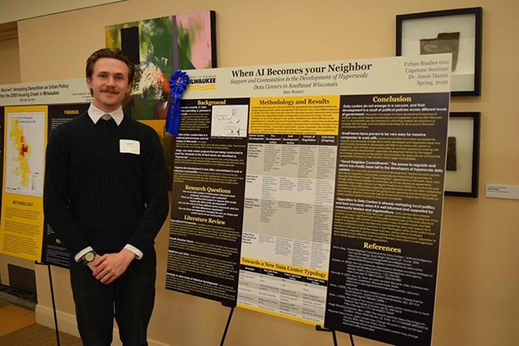 A young white man with short brown hair and a mustache dressed in business casual stands next to a black, white, and yellow research poster. A young white man with short brown hair and a mustache dressed in business casual stands next to a black, white, and yellow research poster.