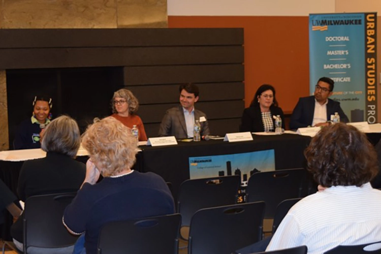 A diverse group of five people sit at a table in front of a small audience. The table has a UWM Urban Studies banner hanging from the edge. A diverse group of five people sit at a table in front of a small audience. The table has a UWM Urban Studies banner hanging from the edge.