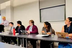 A line of six adults sit at a long gray table, some with laptops in front of them.