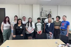 A group of nine students and their faculty advisor stand in a row behind a table smiling at the camera. Behind them is a shelf holding several tall trophies.