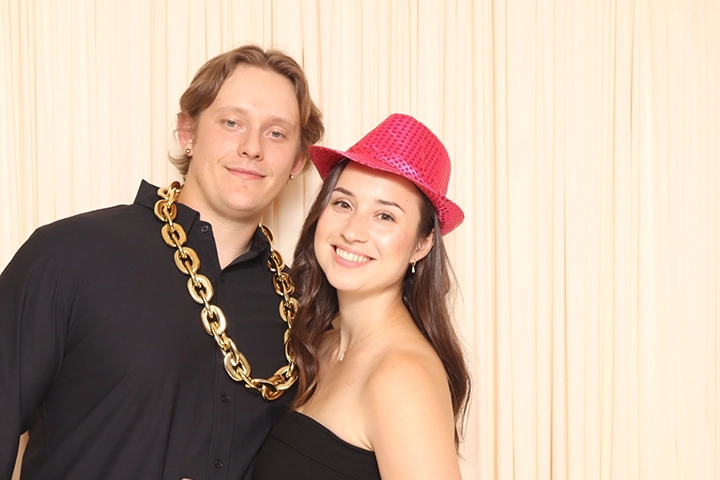 A young white man and woman stand together in front of a cream-colored drape background. They both wear black shirts; the man has a fake gold chain around his neck and the woman wears a sparkly red hat.