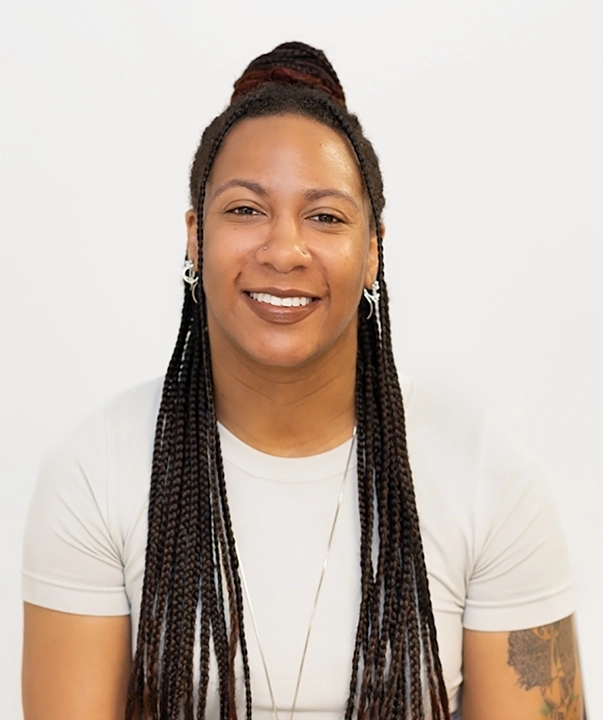 A headshot of a Black woman with long braided hair. She wears a white t-shirt and has silver earrings.