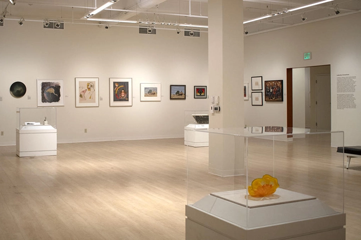 An open airy room with wood floors and white walls. Several works of art hang on the walls. Various display cases with 3D art are around the room.