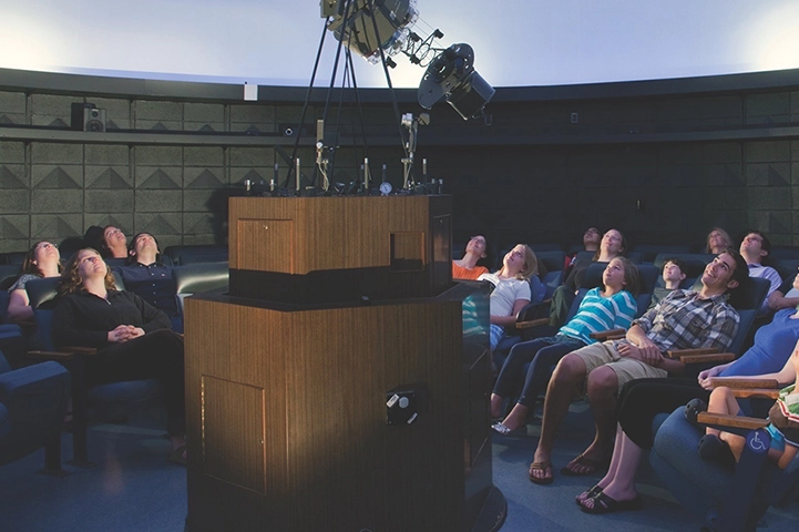 A crowd of people sit in chairs arranged in a circle around a star projector. They all look up at the white domed ceiling.