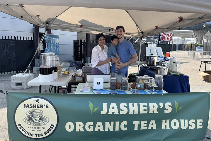 A southeast Asian woman and a white man stand holding their child between them. They are behind a table labeled with a banner for Jasher's Organic Tea House. Vats of tea sit oun tables around them beneath a tent.