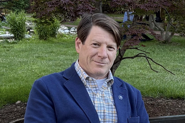 Headshot of a middle-aged white man with short brown hair wearing a collared shirt and blue blazer. In the background are trees and a grassy field.